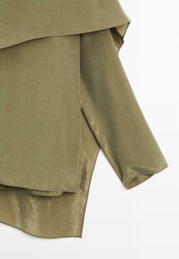 Vêtement superposé vert olive avec une texture lisse, présentant un design drapé et un ourlet incurvé. Tissu doux avec un léger éclat.