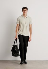 Polo-shirt beige clair à manches courtes, associé à un pantalon noir. Portant un tote bag noir. Design simple et moderne.