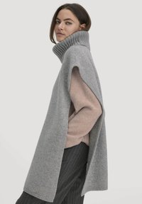 Grijze gebreide oversized trui met een hoge ribbelkraag, zijsplitten en een zachte textuur, gelaagd over een roze kledingstuk en gestreepte broek.