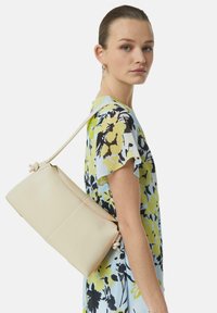 Kremowa skórzana torba o gładkiej fakturze, zaokrąglonym kształcie i regulowanym pasku noszona na ramieniu. Posiada minimalistyczne detale szycia.