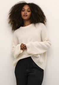Krémový oversized svetr s kulatým výstřihem a měkkou texturou, kombinovaný s černými, texturovanými a zúženými kalhotami.
