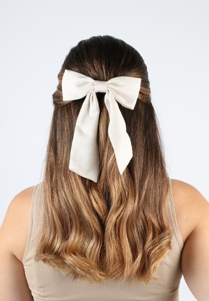 Cremefarbener Seidenbogen mit langen Enden, der im hinteren Teil von welligem, braunem Haar befestigt ist. Der Hintergrund ist schlicht weiß, was das Haaraccessoire hervorhebt.