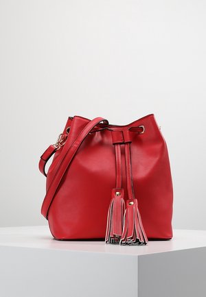Rode leren handtas in emmerstijl met verstelbare schouderriem en twee kwastjes die aan de voorkant hangen, geplaatst op een wit oppervlak.