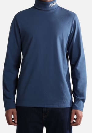 Mężczyzna ubrany w granatowy golf z długim rękawem z logo "Napapijri" na kołnierzu, w połączeniu z ciemnymi jeansami.