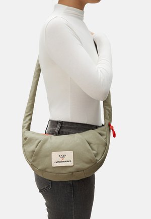 Person trägt ein weißes Langarmshirt und dunkle Jeans und trägt eine gepolsterte olivgrüne Umhängetasche mit sichtbarem Etikett und rotem Reißverschluss.