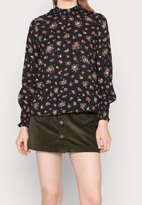 Donna che indossa un blouse nero a maniche lunghe con stampa floreale rosa e una gonna mini in velluto a coste verde scuro con bottoni.