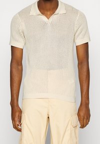 Beżowa dzianinowa koszulka polo z teksturowanym wzorem, krótkimi rękawami i dekoltem w serek. Tkanina jest przewiewna.