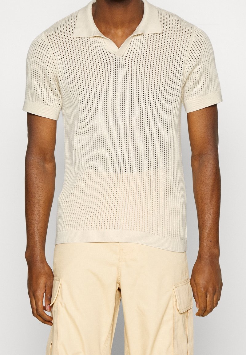 Beżowa dzianinowa koszulka polo z teksturowanym wzorem, krótkimi rękawami i dekoltem w serek. Tkanina jest przewiewna.
