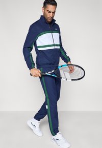 Chándal azul marino con franjas blancas y verdes, chaqueta con cremallera, puños elásticos y pantalones a juego. Raqueta de tenis en la mano derecha. Zapatillas blancas.