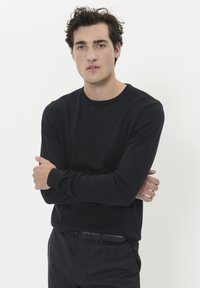 Svart långärmad tröja med rund halsringning i ett mjukt tyg, bärs med grå mönstrade byxor och ett svart bälte, modellen poserar med armarna i kors.