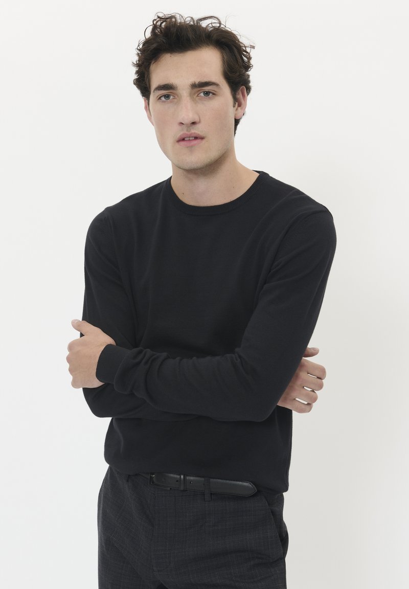 Svart långärmad tröja med rund halsringning i ett mjukt tyg, bärs med grå mönstrade byxor och ett svart bälte, modellen poserar med armarna i kors.