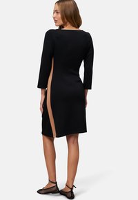 Zwarte jurk met een aansluitende silhouet, driekwart mouwen en een hoge split aan de zijkant met een beige paneel. Glad stofoppervlak.