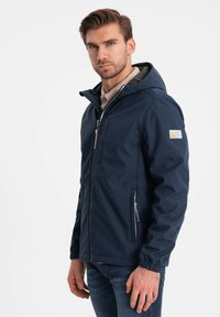 Marineblå softshell-jakke med lynlås foran, justerbar hætte, elastiske manchetter og sidelommer med lynlås.