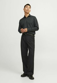 Camisa negra de botón con un diseño elegante, combinada con pantalones oscuros y un cinturón texturizado; los zapatos son negros brillantes, completando el look profesional.