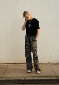 Black t-shirt with a graphic design, patterned black and white pants with geometric shapes, white footwear, standing against a textured wall.