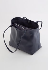 Bolso tote de cuero azul marino con una textura suave, dos asas y un diseño de apertura en la parte superior. El interior cuenta con un bolsillo con cremallera para un almacenamiento seguro.