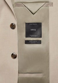 Interno della giacca beige con due bottoni marroni, etichetta nera del marchio e cartellino super slim fit. Tessuto liscio con texture e dettagli minimi.