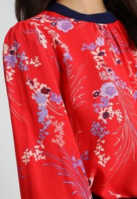Rote Bluse mit floralen Mustern und navyfarbenem Kragen. Mit Puffärmeln und einem leichten, glatten Stoff, der mit lila und weißen Blumenmustern verziert ist.