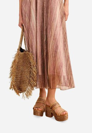 Mujer que lleva una larga falda plisada rosa y dorada, sosteniendo un gran bolso de paja tejido y calzando sandalias altas de plataforma tejidas.