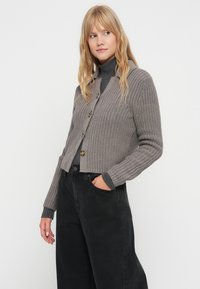 Siv prepleten kardigan z vzorom, okroglim ovratnikom in zapenjanjem na gumb, v kombinaciji z črnimi širokimi kavbojkami in sivim puloverjem s turtleneck ovratnikom.