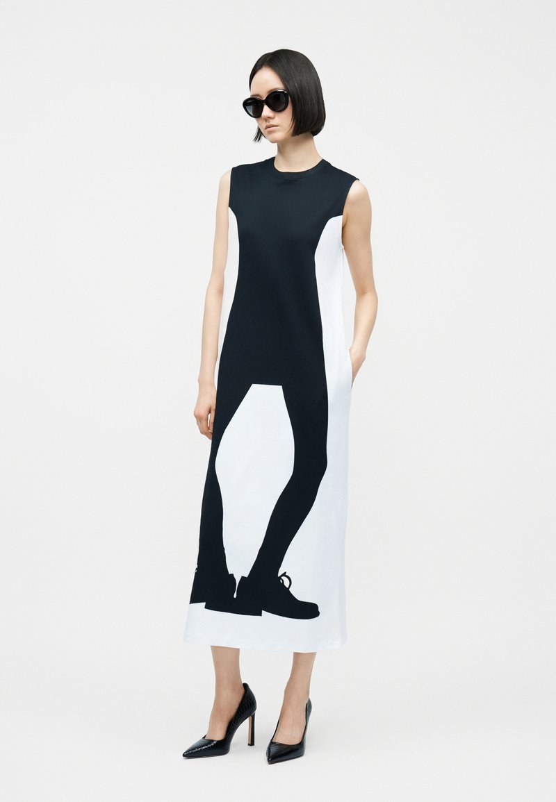Femme portant une robe midi sans manches noire et blanche avec un imprimé silhouette noire audacieux et des talons hauts noirs, posant avec une main dans la poche.