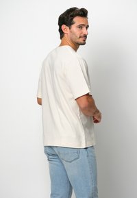 Beżowa koszulka krótki rękaw wykonana z bawełny, o luźnym kroju, z prostym okrągłym dekoltem i prostym dołem, w połączeniu ze jasnoniebieskimi dżinsami.