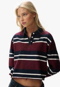 Ung kvinde med blond hår iført en kortærmet rugbytrøje med lange ærmer, der har maroon, navy og hvide vandrette striber.