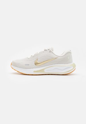 Chaussure de course Nike blanche et beige avec logo swoosh doré, empeigne texturée, semelle rembourrée et semelle extérieure orange vue de côté.