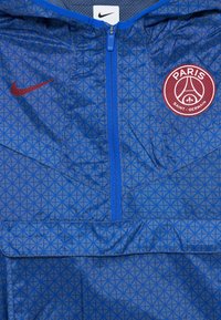 Veste de sport bleue avec un logo Nike rouge sur la gauche et le badge du Paris Saint-Germain sur la droite, dotée d'une fermeture éclair frontale et d'un motif géométrique.