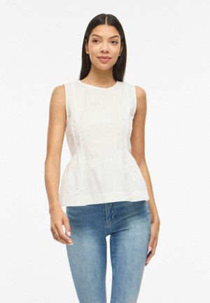 Kvinne med rett mørkt hår iført ermeløs hvit bluse med folder og blå jeans, stående mot hvit bakgrunn.