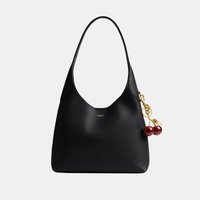 Bolso hobo de cuero negro con una sola correa, pequeño logo dorado de Coach en el centro y un colgante dorado con forma de cereza colgando al lado.