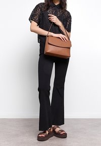 Femme en blouse en dentelle noire et pantalon tenant un sac à bandoulière en cuir marron, portant des sandales à plateforme avec des lanières de marque.