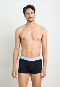 Black boxer shorts with a light blue waistband featuring the "LACOSTE" logo. The material appears soft and fitted. Model stands against a neutral background.