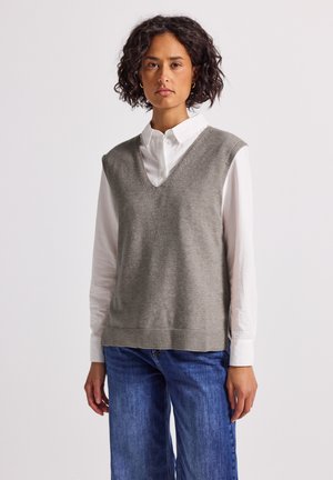 Kvinna med lockigt hår som bär en vit knapplös skjorta, grå ärmlös väst och blå jeans mot en helt vit bakgrund.