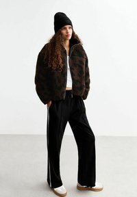 Bruin en zwart luipaardprint fleece jack, met rits, gecombineerd met zwarte wijde broek en crèmekleurige sneakers met bruine zolen. Zwarte beanie.