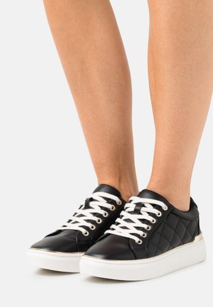 Piernas con zapatillas negras acolchadas con cordones blancos y suelas gruesas blancas sobre un fondo gris claro.