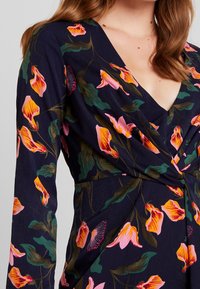 Sukienka w stylu wrap z kwiatowym wzorem w granatowym kolorze, zdobiona różowymi i żółtymi kwiatami, z długimi rękawami, głębokim dekoltem w kształcie litery V oraz teksturowanym wykończeniem.