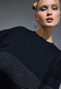 Noor naine, kellel on sinine silmavärv, rõngakujulised kõrvarõngad ja punutud juuksed, kannab musta topi, mis on kaunistatud väikeste hõbedaste naelte poolkaarega.