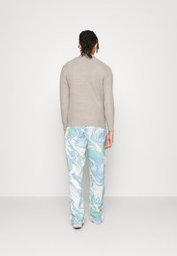 Man med flätat hår som bär en beige stickad tröja, byxor med flerfärgad blå-grön virvelformad mönstring och vita sneakers, vänd bort från kameran mot en enkel bakgrund.