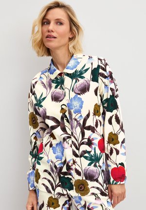 Vrouw met kort blond haar draagt een overhemd met knopen met grote kleurrijke bloemen- en bladmotieven op een lichte achtergrond.