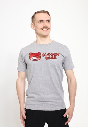 Jonge man met kort haar en snor draagt een grijs T-shirt met een rood "Gloomy Bear" logo en een cartoon berengezicht.