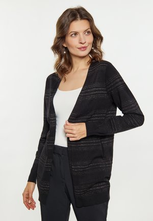 Frau mit schulterlangem, welligem Haar, die Perlenohrringe trägt, eine schwarze offene Strickjacke mit dezenten Streifen, weißes Oberteil und schwarze Hose.