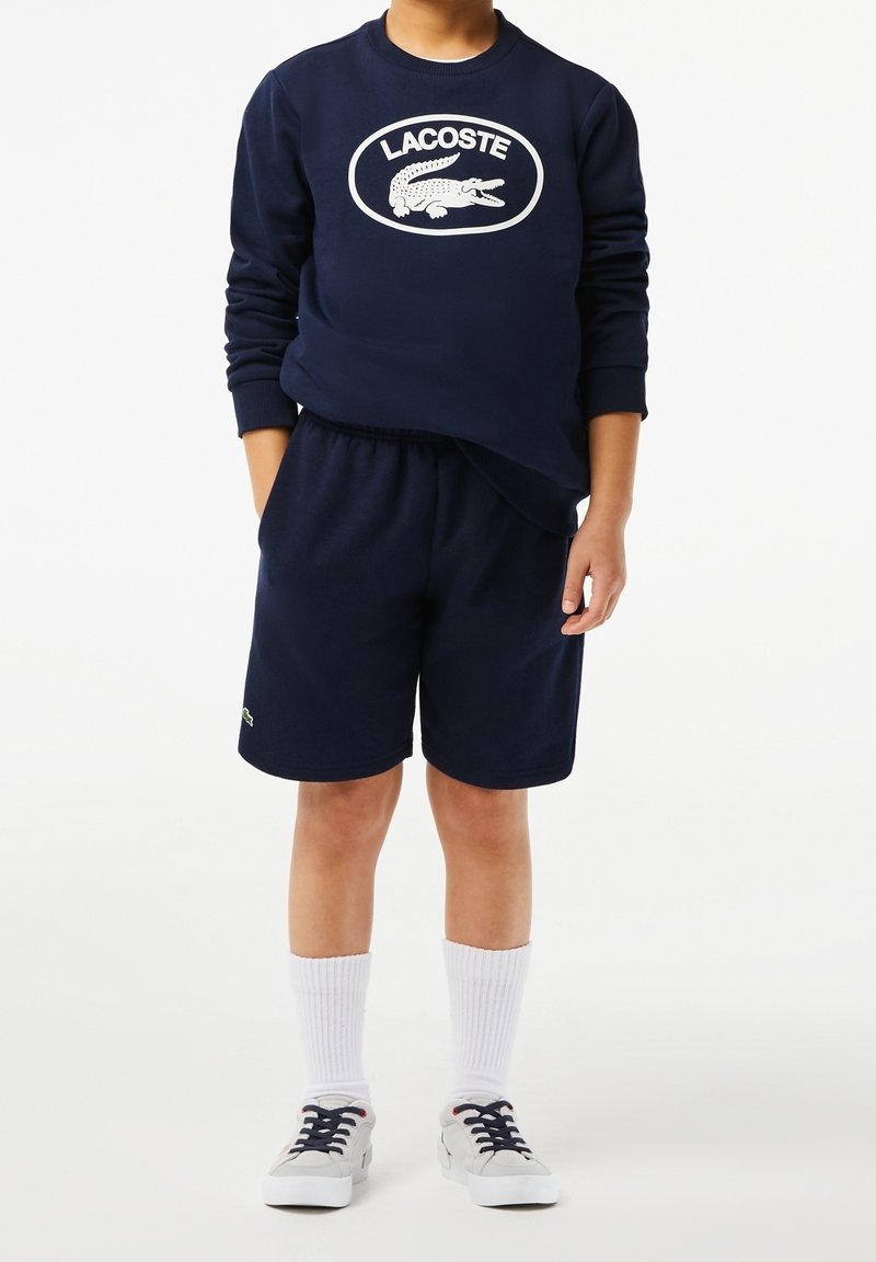 Enfant portant un sweat-shirt Lacoste bleu marine avec logo crocodile, short assorti, chaussettes blanches mi-mollet et baskets blanches, debout devant un fond blanc.