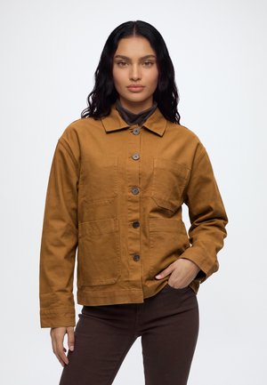 Une femme porte une veste brun moutarde boutonnée sur le devant et dotée de grandes poches, associée à un pantalon foncé sur un fond clair uni.
