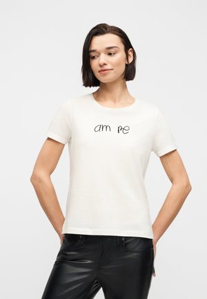 Donna con capelli corti e scuri che indossa una maglietta bianca con la scritta "am re" e pantaloni in pelle neri, in piedi con le mani dietro la schiena.