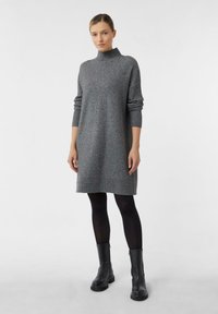 Frau in einem grauen Langarm-Strickkleid, schwarzen Strumpfhosen und schwarzen Knöchelstiefeln, steht vor einem schlichten weißen Hintergrund.