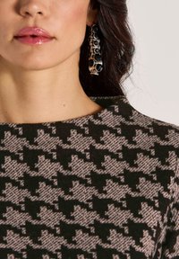 Vrouw met glanzende roze lippen, draagt grote zilveren oorbellen en een zwart-beige houndstooth patroon top met een hoge hals.