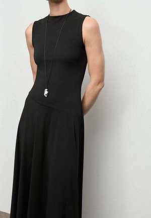 Ärmelloses schwarzes Kleid mit tailliertem Oberteil und fließendem Rock, getragen mit einer langen Halskette mit silbernem Anhänger, vor einfachem hellem Hintergrund.