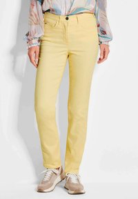 Gelbe Stretch-Jeans im Slim-Fit-Stil, mit einem vorderen Knopfverschluss, fünf Taschen und kontrastierendem Nähen. Tragen mit beigen Sneakers.