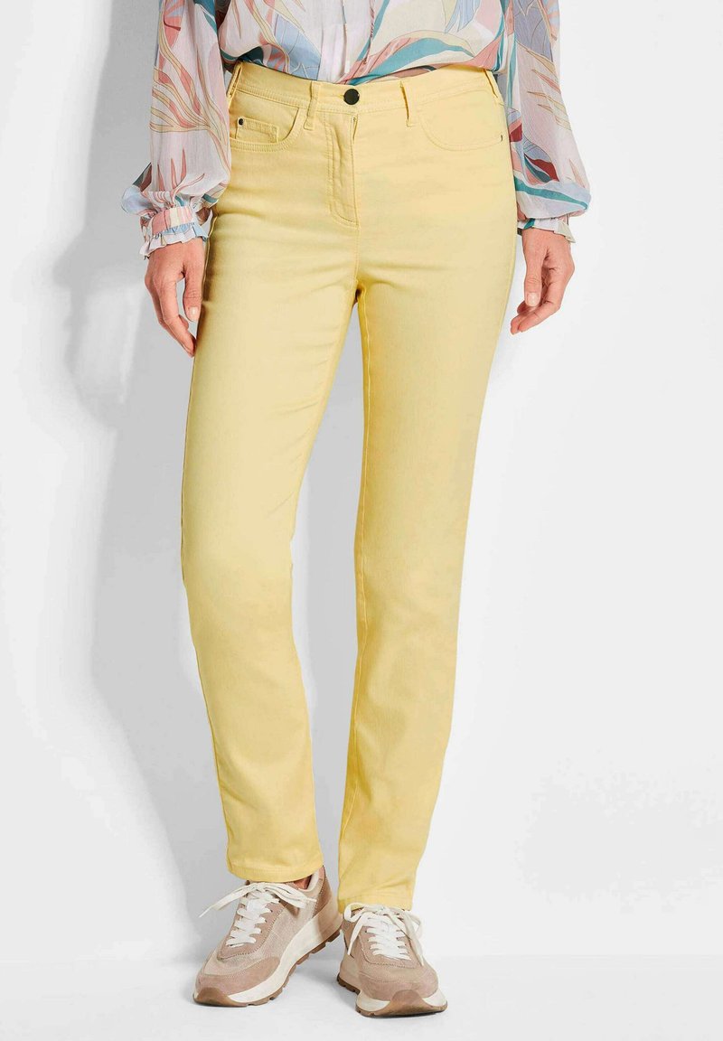 Gelbe Stretch-Jeans im Slim-Fit-Stil, mit einem vorderen Knopfverschluss, fünf Taschen und kontrastierendem Nähen. Tragen mit beigen Sneakers.